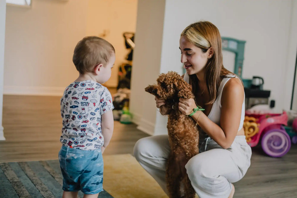 A young woman crouching and holding a brown poodle, with a toddler looking on.
